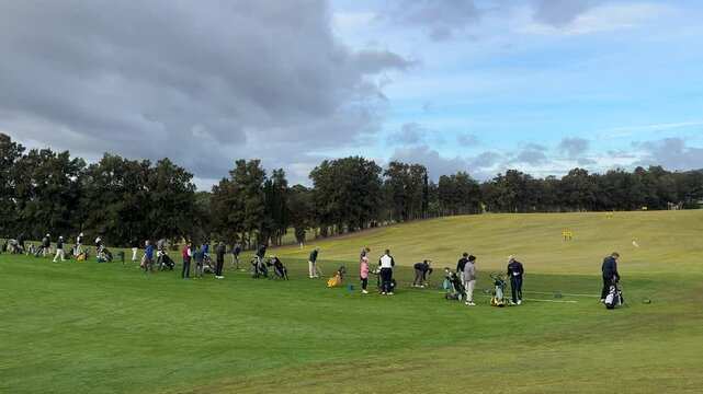 Golfers practicing their swings on a driving range surrounded by trees under a partly cloudy sky.
