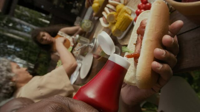 Close-up rolling shot of middle-aged African American man picking up bottle of ketchup, adding tomato sauce on top of hot dog sausage while enjoying outdoor picnic with family on summer day