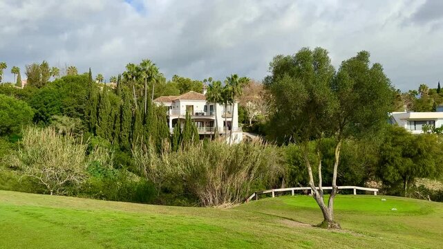 Mediterranean villa surrounded by lush greenery and palm trees, viewed across a manicured lawn on a bright day with scattered clouds in Sotogrande, Spain.
