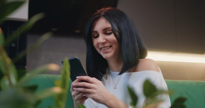 Female recording audio message for friends or parents, sharing news using mobile phone application. Pretty woman sit in kitchen hold smart phone talk on speakerphone.