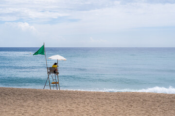 A solitary figure vigilantly surveys the tranquil waters from an elevated chair on a peaceful beach