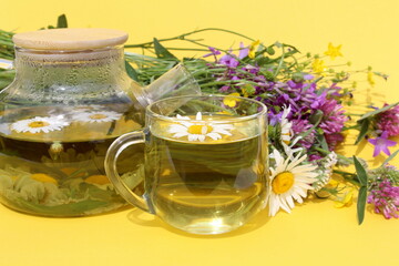 On a yellow background there is a glass teapot with a cup of chamomile infusion.	