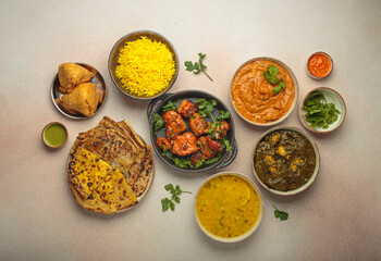 Overhead view of a variety of Indian dishes on a beige table, including curry, tandoori chicken, chicken tikka masala, dal, Palak paneer, paratha, rice, samosas, rich culinary heritage of India