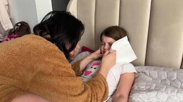 Mother tries to pull a loose tooth from her daughter who resists and cries in bed, expressing fear and discomfort.
