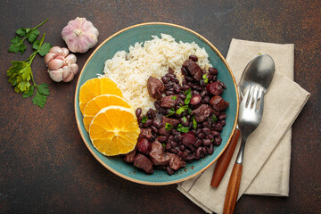Hearty brazilian feijoada stew with black beans, pork, beef, rice, and orange slices in rustic bowl on wooden board with cutlery and napkin. Traditional dish of Brazil