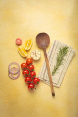 Cherry tomatoes, garlic, lemon slices, carrots, red onions, and rosemary sprig arranged alongside a wooden spoon on a yellow backdrop, culinary creativity and healthy eating concept
