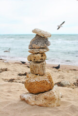 A carefully balanced stack of stones stands on the sandy beach, with gentle ocean waves in the background. A seagull flies overhead, enhancing the serene coastal atmosphere
