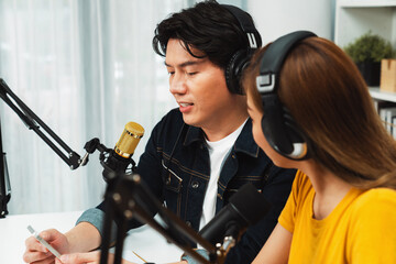 Smiling radio influencer hosts wearing headphones with channel social media online live streaming with talking topic on script note and laptop to listeners at morning time at studio record. Infobahn.