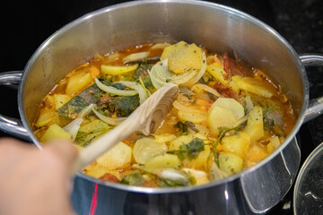 Traditional Portuguese codfish stew, known as caldeirada de bacalhau, prepared in a stainless steel pot with sliced potatoes, onions, tomatoes, peppers, herbs and olive oil.Homemade meal on a stovetop