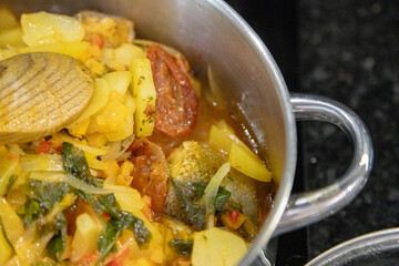 Traditional Portuguese codfish stew, known as caldeirada de bacalhau, prepared in a stainless steel pot with sliced potatoes, onions, tomatoes, peppers, herbs and olive oil.Homemade meal on a stovetop