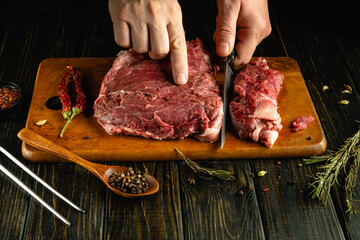 Skilled hands slice a raw piece of meat on a wooden cutting board in a rustic kitchen. Dried herbs and spices surround the preparation area, enhancing the culinary atmosphere