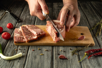 Chef slices a thick piece of meat with a sharp knife on a rustic wooden cutting board surrounded by...