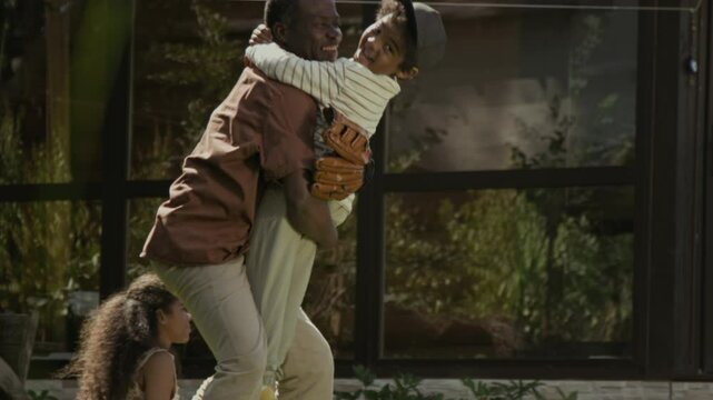 Wide shot of black dad training teenage son in baseball skills on green lawn of suburban family home, then hugging little boy running towards him. Family relaxing together on hot summer day