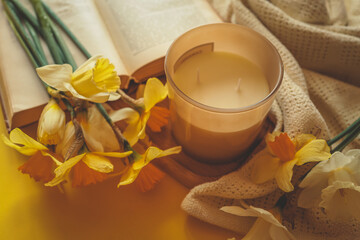 Unlit candle surrounded by yellow daffodils, a soft knitted fabric, and an open book, creating a warm and peaceful spring scene with cozy natural tones
