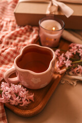 A cozy spring scene with a pink floral cup of tea, a lit candle, pink hyacinths, and a gift box on a wooden tray with soft fabrics in warm tones