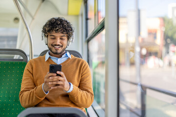 A smiling man uses his phone while riding on a bus, wearing headphones and an orange sweater