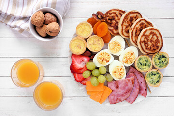 Breakfast or brunch grazing board. Collection of items including fruit, ham, quiche, pancakes and eggs. Top view table scene on a white wood background.