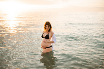 Pregnant woman wearing black swimsuit in sea water over sunset outdoors. Summer vacation time. Maternity.