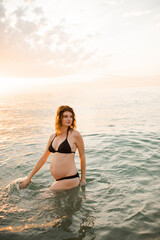 Pregnant woman wearing black swimsuit in sea water over sunset outdoors. Summer vacation time. Maternity.