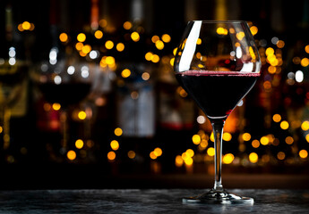 Red merlot wine glass and wine bottles on dark bar background with lights