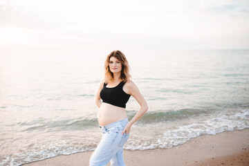 Pregnant woman wearing denim pants and black top over sea and sunset at beach outdoors. Maternity clothes. Summer vacation season.
