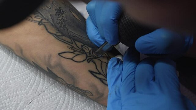 Close-up view of a tattoo artist in blue gloves creating a floral tattoo on a clients forearm. The tattoo machine is in motion as the artist outlines the design. Studio environment