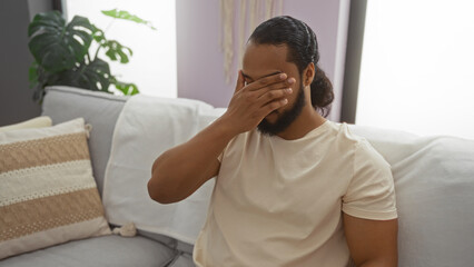 Young man with beard sitting indoors covering face in cozy living room with pillows and houseplants suggesting contemplation or stress in an interior home setting