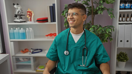 Young man in a clinic room wearing teal scrubs and stethoscope, smiling with medical equipment around, suggesting a healthcare setting.