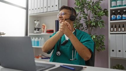 Fototapeta premium Young male healthcare professional in clinic setting smiling while using laptop with headset, surrounded by medical equipment and office files.