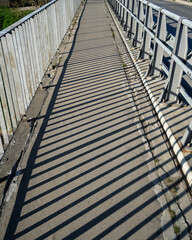 Rhythmic pattern of long parallel shadows cast by railings across a sunlit pedestrian bridge walkway. Strong light and shadow interplay emphasizing lines perspective and structure