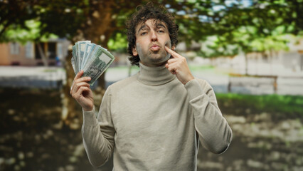 Fototapeta premium Hispanic man holding bangladesh taka banknotes in an outdoor park setting, expressing thoughtfulness amid lush greenery and a blurred urban background.