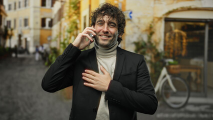 Young hispanic man in city street smiling with one hand on chest while talking on phone, wearing casual jacket and surrounded by urban architecture and bicycles.
