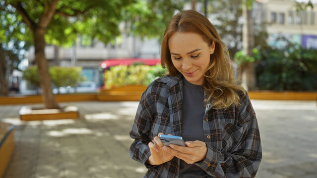 Woman smiling while using smartphone in city park surrounded by urban scenery and greenery on sunny day blending nature and modern life outdoors