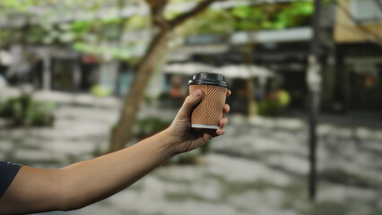 Man holding a coffee cup on a city street with a blurred outdoor background of buildings and trees.
