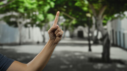 Man's hand pointing with index finger while standing on an urban street lined with green trees,...