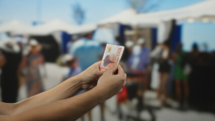 Man holding norwegian krone banknote outdoors in a vibrant market street amidst busy shoppers and shops under sunlit tents.