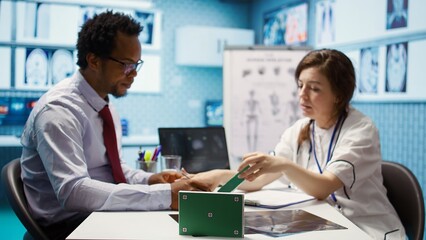 Obraz premium Patient and medical specialist at a check up visit next to green screen on medicine box, discussing about treatment and medical drugs for recovery. Female doctor pointing at medicaments. Camera B.