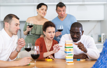 Woman spending free time with group of mates at laid-back home get-together with drinks, concentrating on removing block from tower while playing jenga