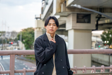A Japanese man in his twenties wearing a black jacket is in Minato Mirai, Yokohama, Kanagawa Prefecture. He sits on a bench, gazing into the distance, surrounded by the calm mood of the modern city.