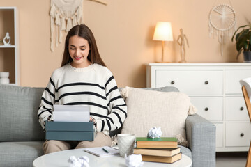 Beautiful happy female writer with typewriter, books and notebook sitting on sofa at home
