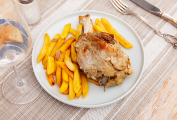 Appetizing spicy baked pork shank with side dish of fried potatoes served on white plate..