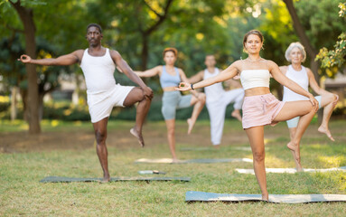 Fit young girl enjoying yoga amidst lush greenery during group class in summer park, performing one...
