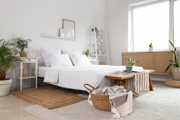 Stylish interior of light bedroom with wooden chest of drawers, bench, houseplants and bed