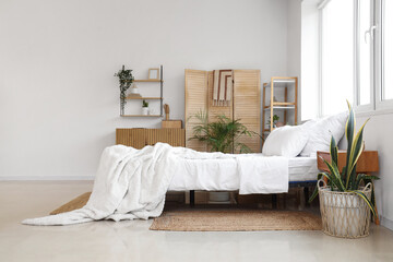 Big bed, shelving unit, folding screen, houseplant and chest of drawers in interior of bedroom