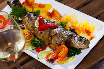 Delicious baked trout served on plate with potatoes, tomatoes, sweet peppers, greens, lemon and white wine