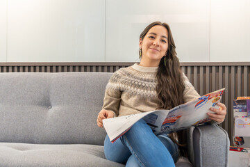 A young woman is sitting comfortably on a modern grey sofa in what appears to be a clinic or office waiting room with a magazine