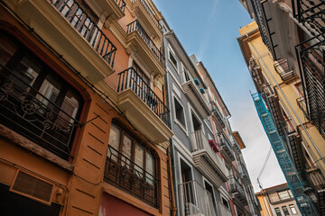 Colourful, balconied apartment blocks crowd a narrow lane in Valencia historic centre, showing wrought‑iron railings, pastel stucco and a slice of blue sky, a symbol of real estate in spain.