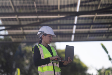 A female technician wearing a safety helmet and reflective vest uses a laptop while inspecting a...