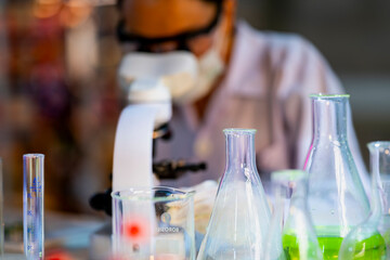 A scientist wearing a mask and gloves works in a lab, examining a green liquid in a beaker and using a microscope. The lab setup is well-lit, reflecting a focus on research, science, and experimentati