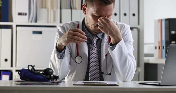 Doctor takes off glasses and rubs nose due to fatigue while sitting at table. Exhausted man demonstrates difficult situation at work in hospital
